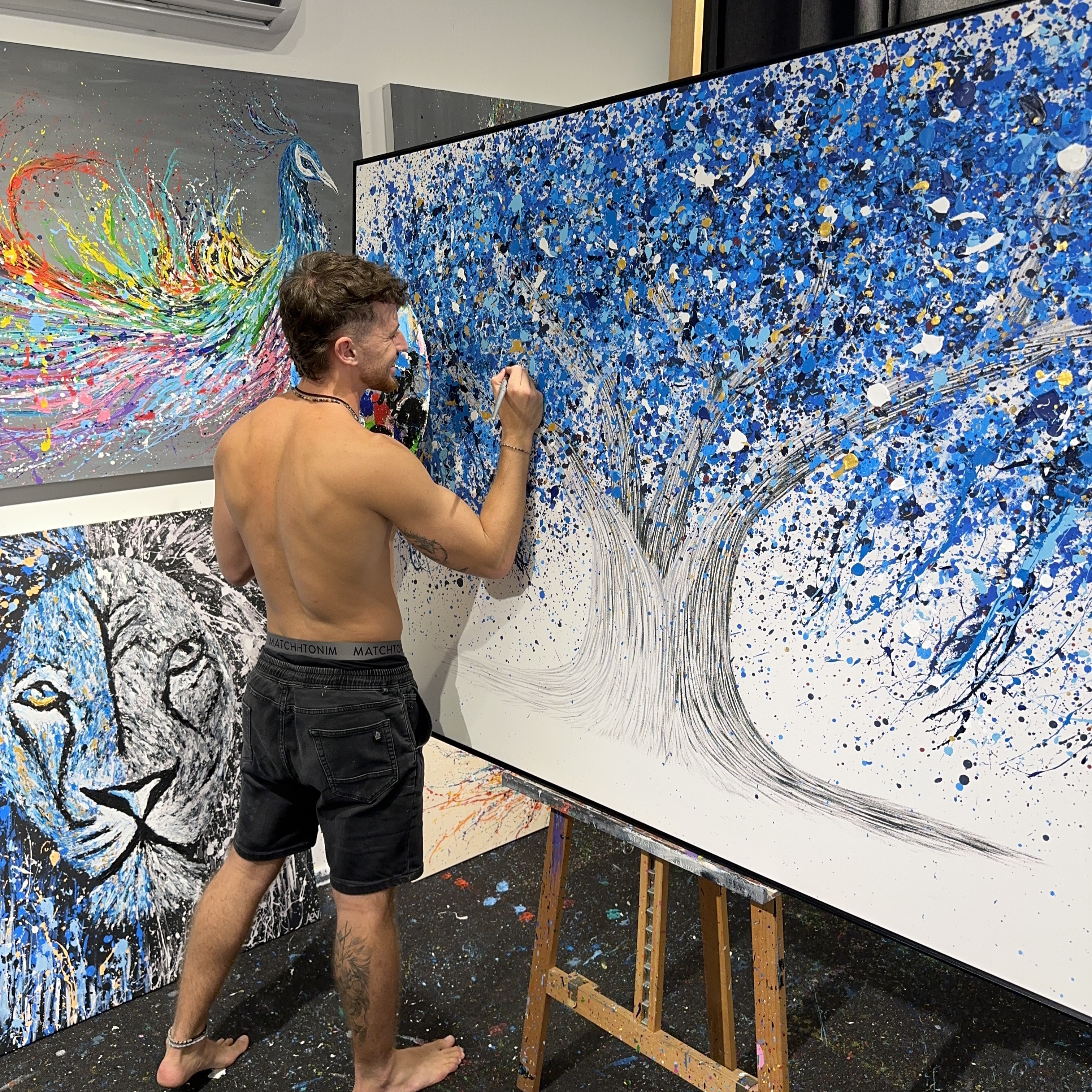 Artist painting a large canvas with a blue tree, surrounded by colorful artworks.