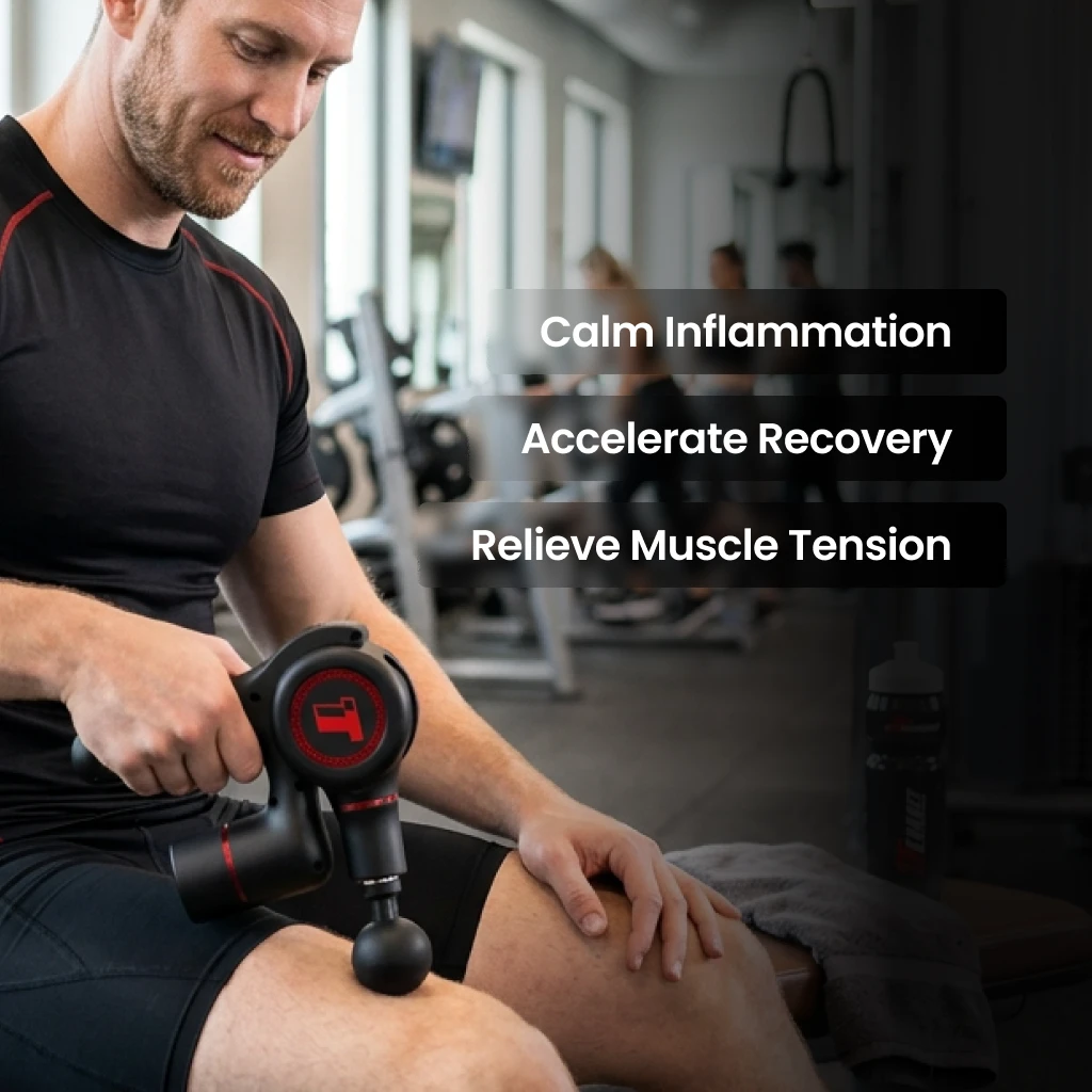 A man in a gym sits on a bench and uses a black and red massage gun on his thigh.