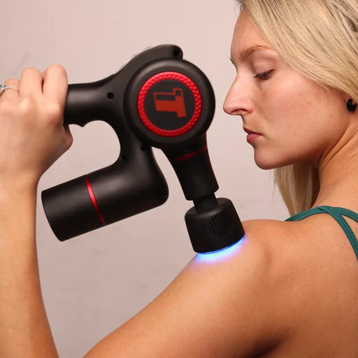 A woman with blonde hair using a black and red percussion massager on her shoulder.