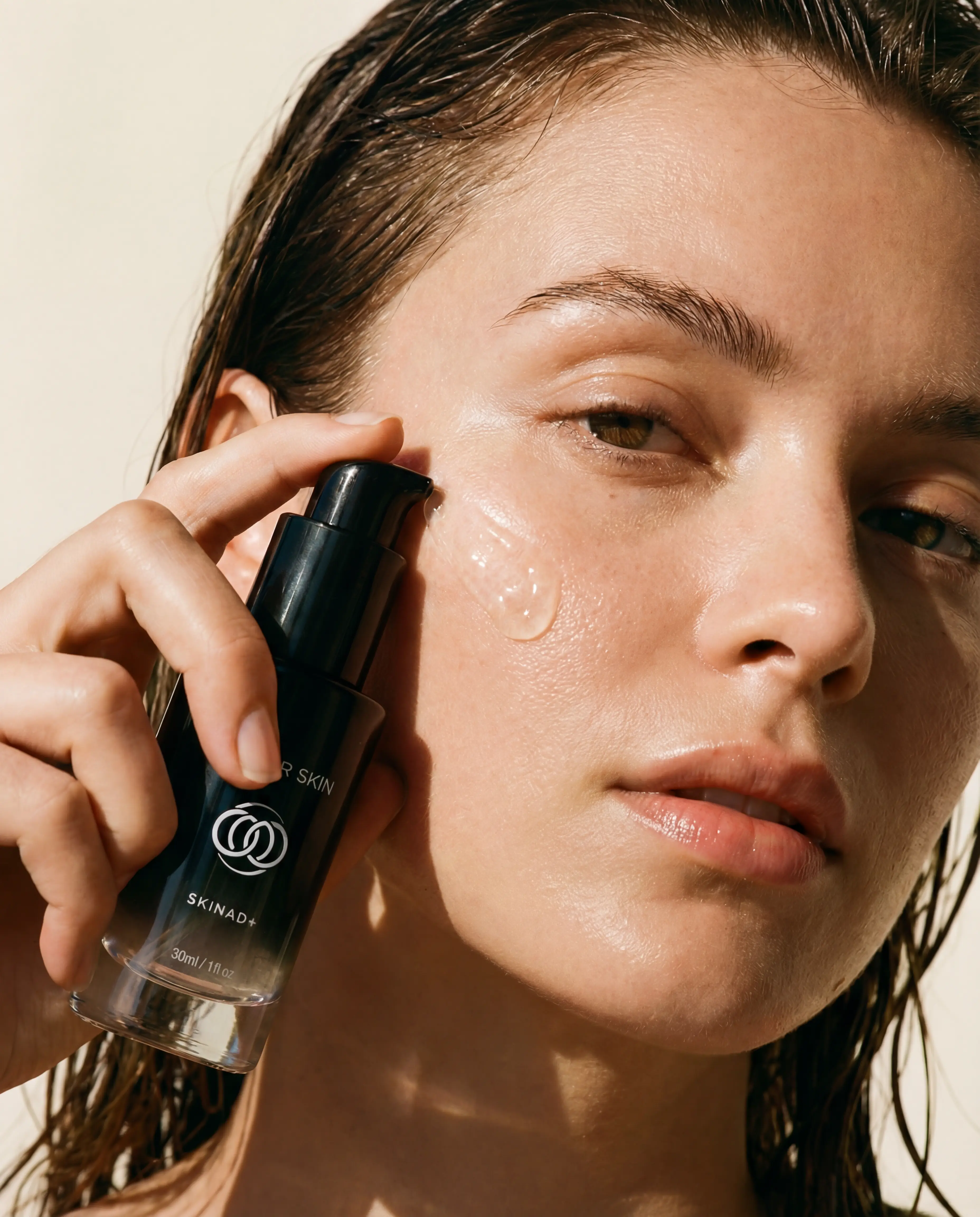 A woman with dewy skin and wet hair applies a clear gel serum to her cheek from a bottle.