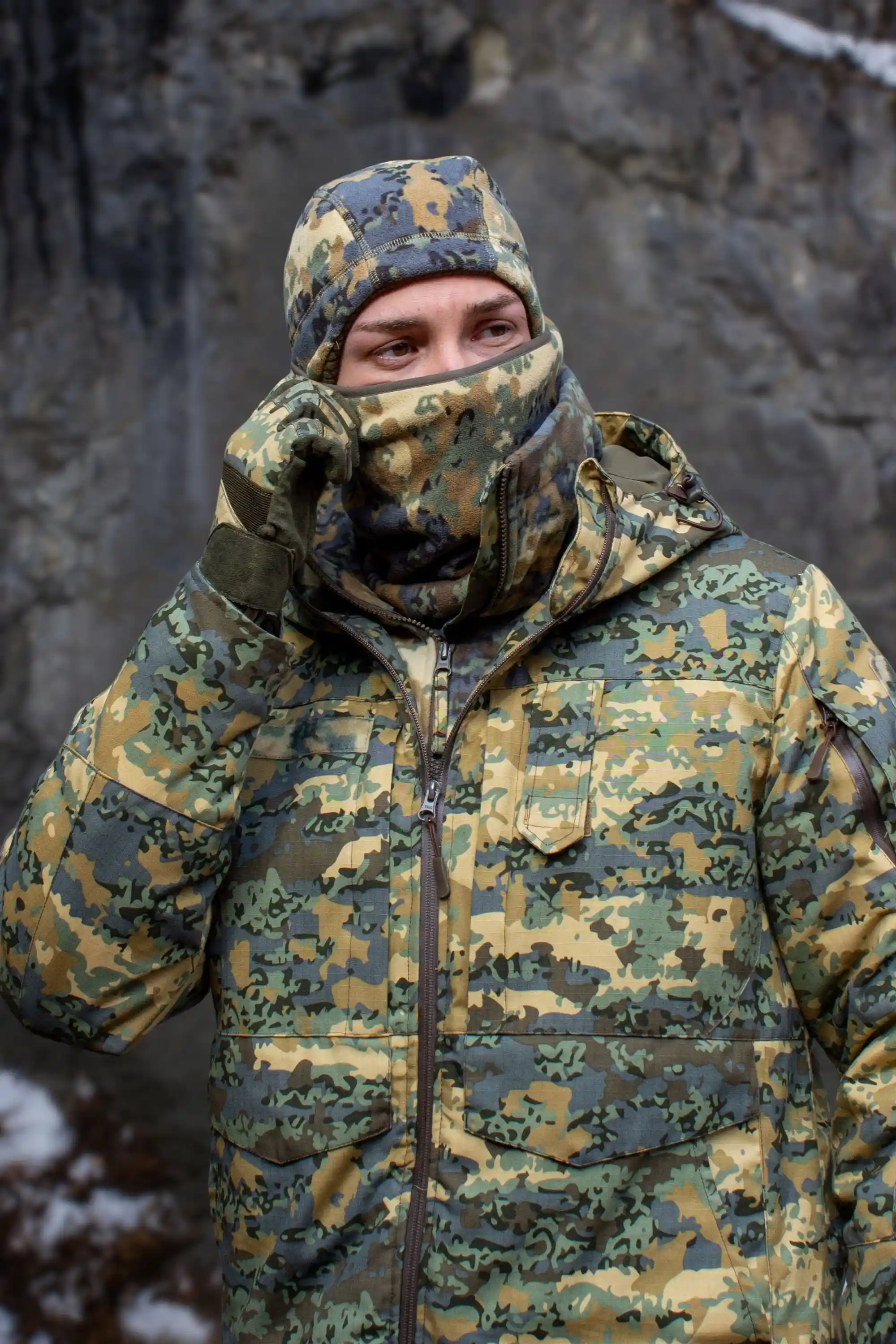 A person in a full camouflage outfit adjusts the balaclava covering their face in front of a rock wall.