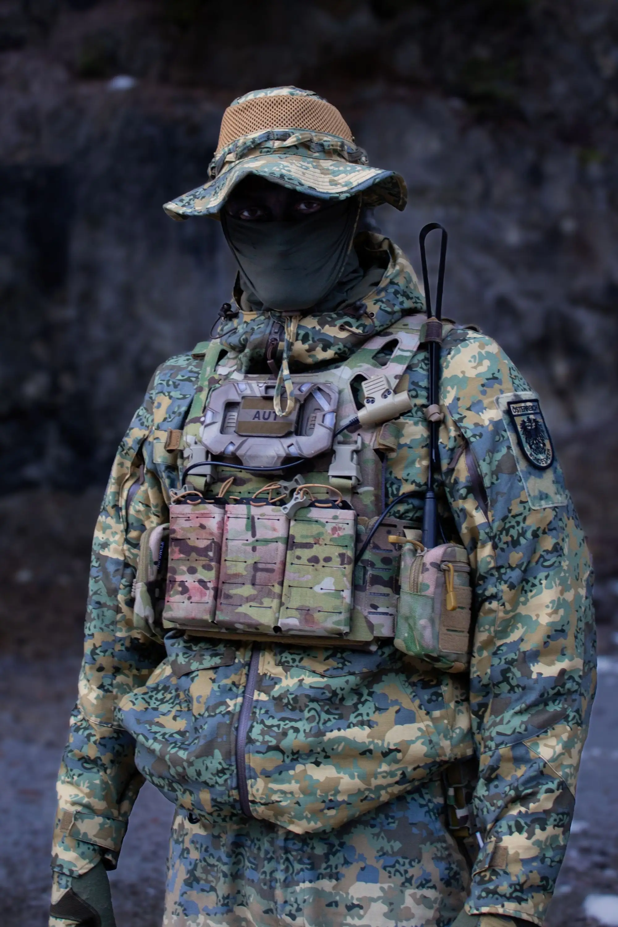 A person in full camouflage military gear, including a vest, boonie hat, and face mask.