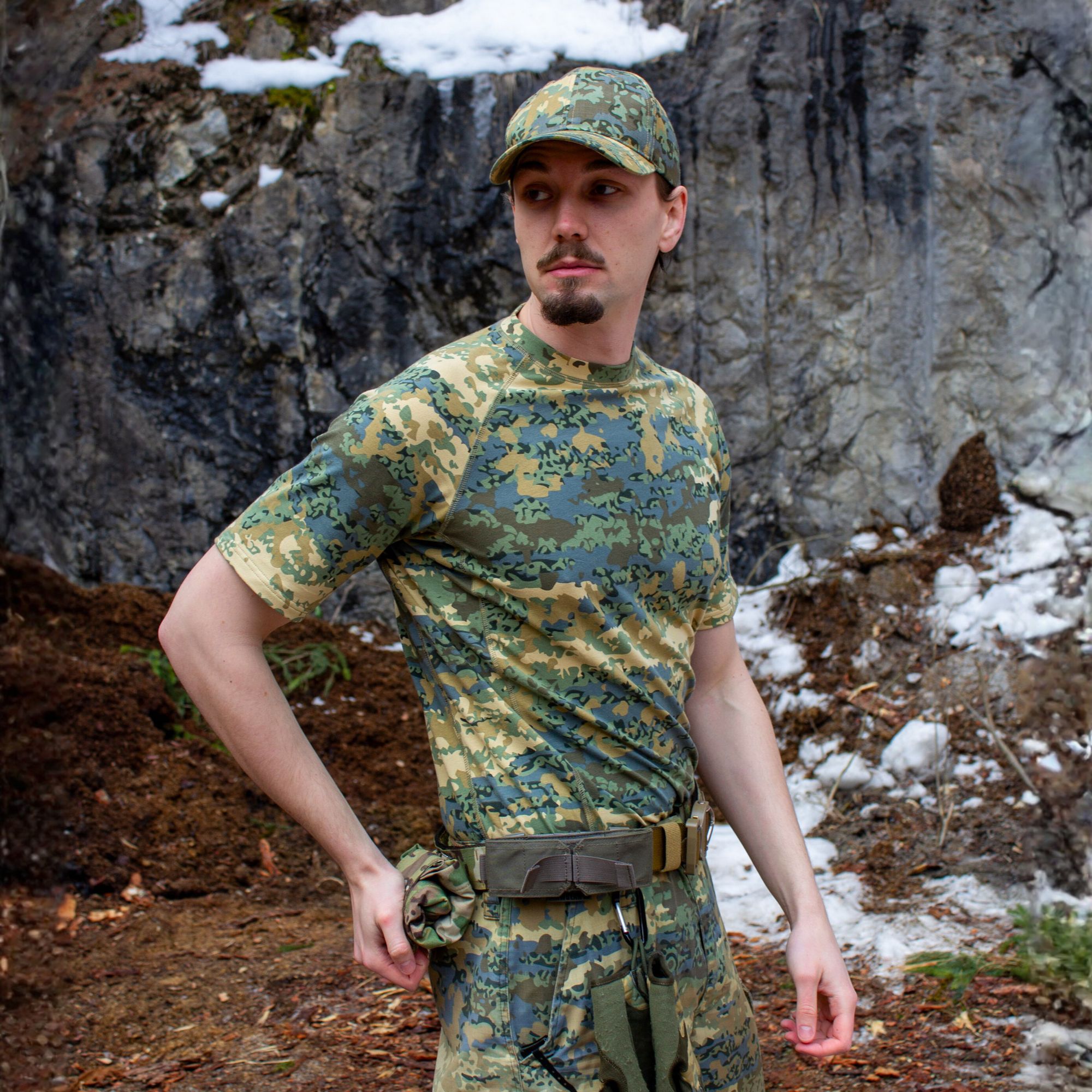 A man in a camouflage t-shirt, pants, and cap stands outdoors in front of a rock face.