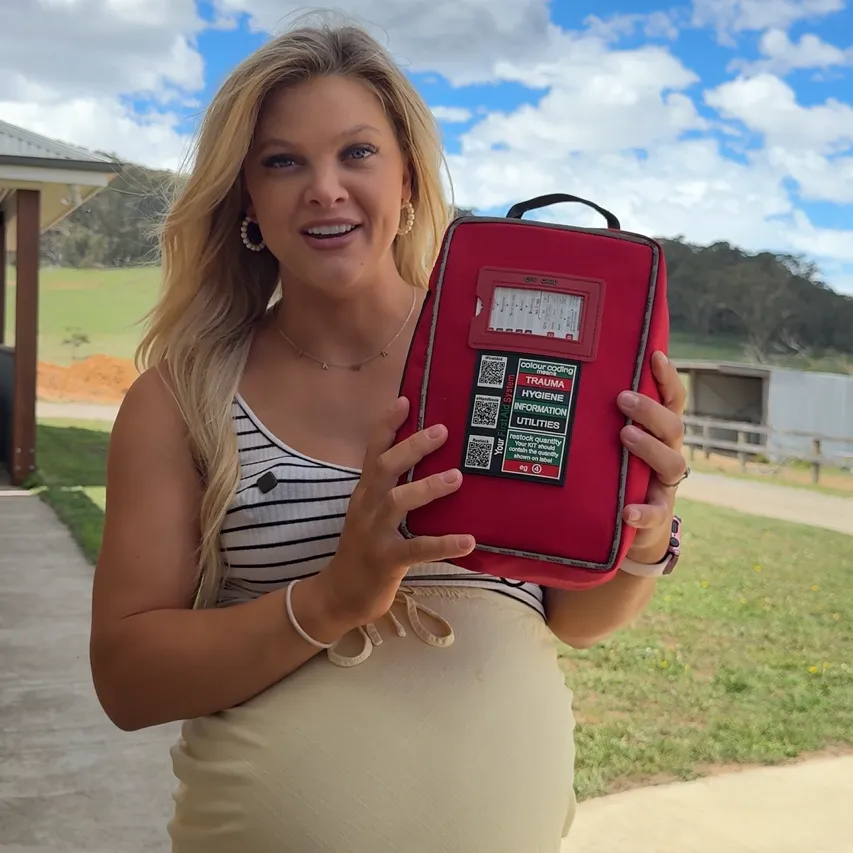 A blonde woman in a striped top stands outside holding a red first-aid kit.