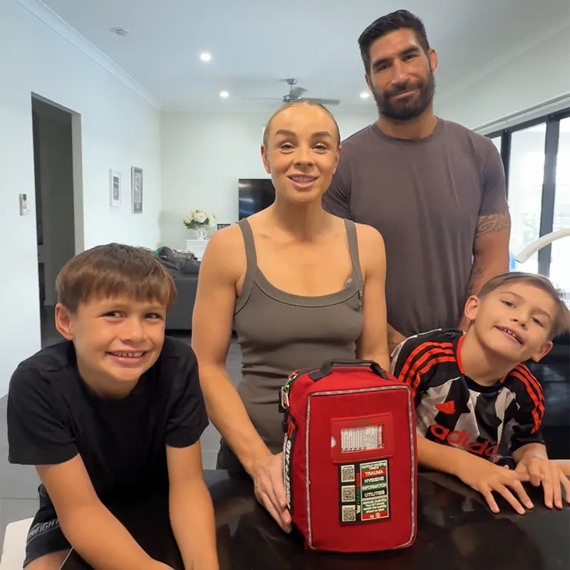 A family of four, two adults and two boys, stand behind a table with a red first-aid kit.