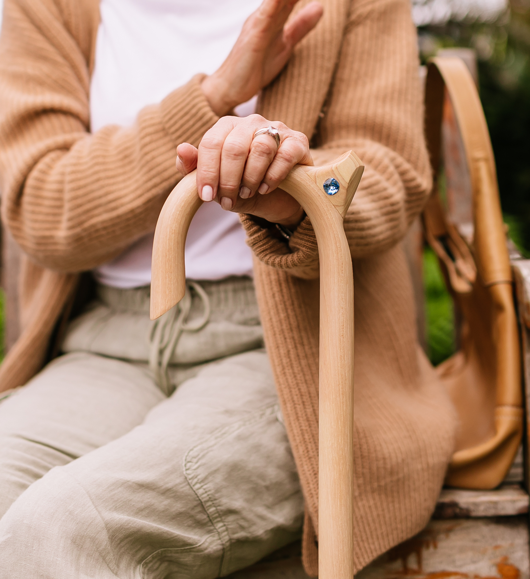 A person's hand resting on a light-colored wooden walking cane with a blue jewel on the handle.