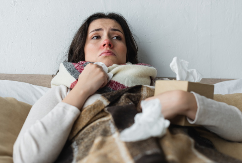 Woman lying in bed with a scarf, holding tissues, looking unwell.
