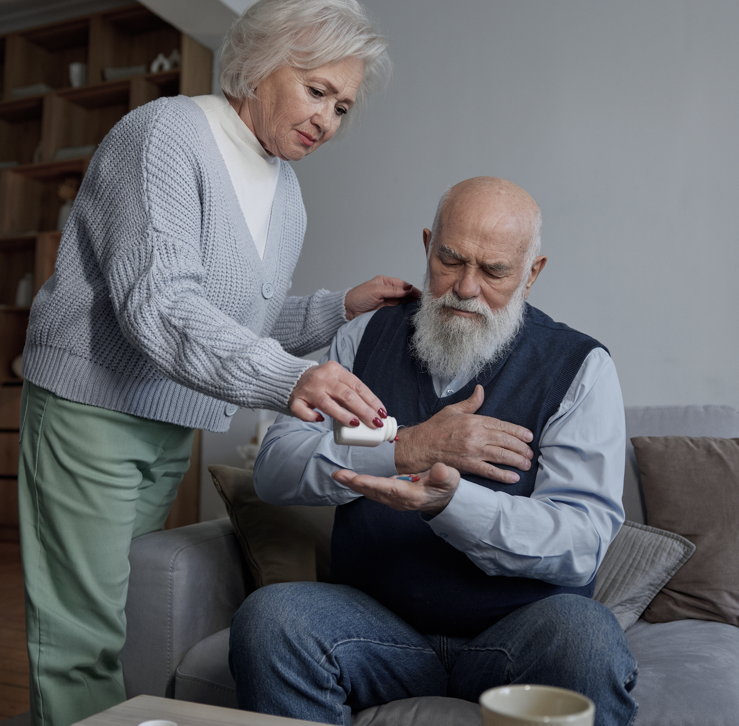 Elderly woman giving pills to an elderly man seated on a sofa.