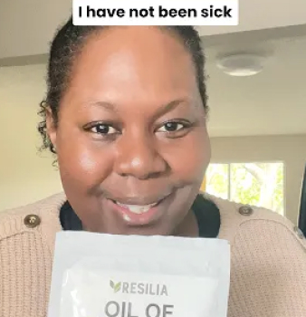 Person smiling, holding a packet labeled 'Resilia Oil of Oregano' indoors.