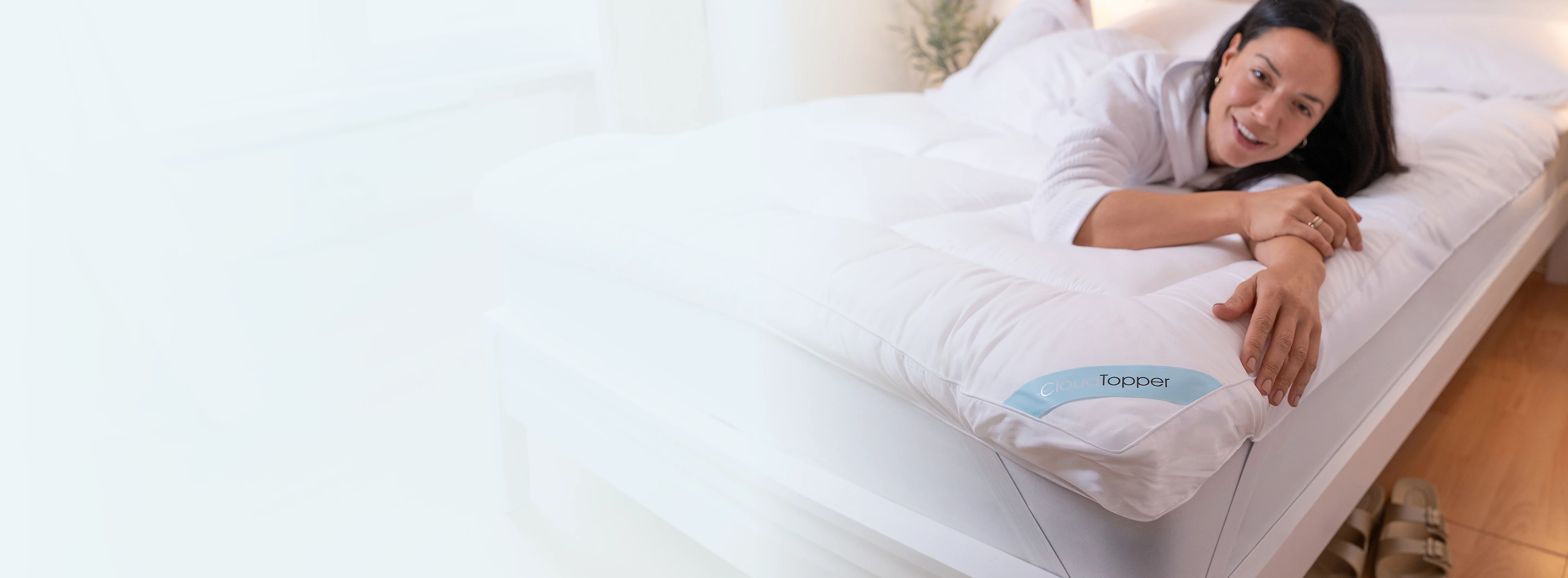 A smiling woman in a white robe lies on a bed with a white mattress topper.