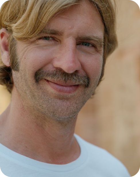 Man with a mustache smiling, wearing a white shirt, blurred background.