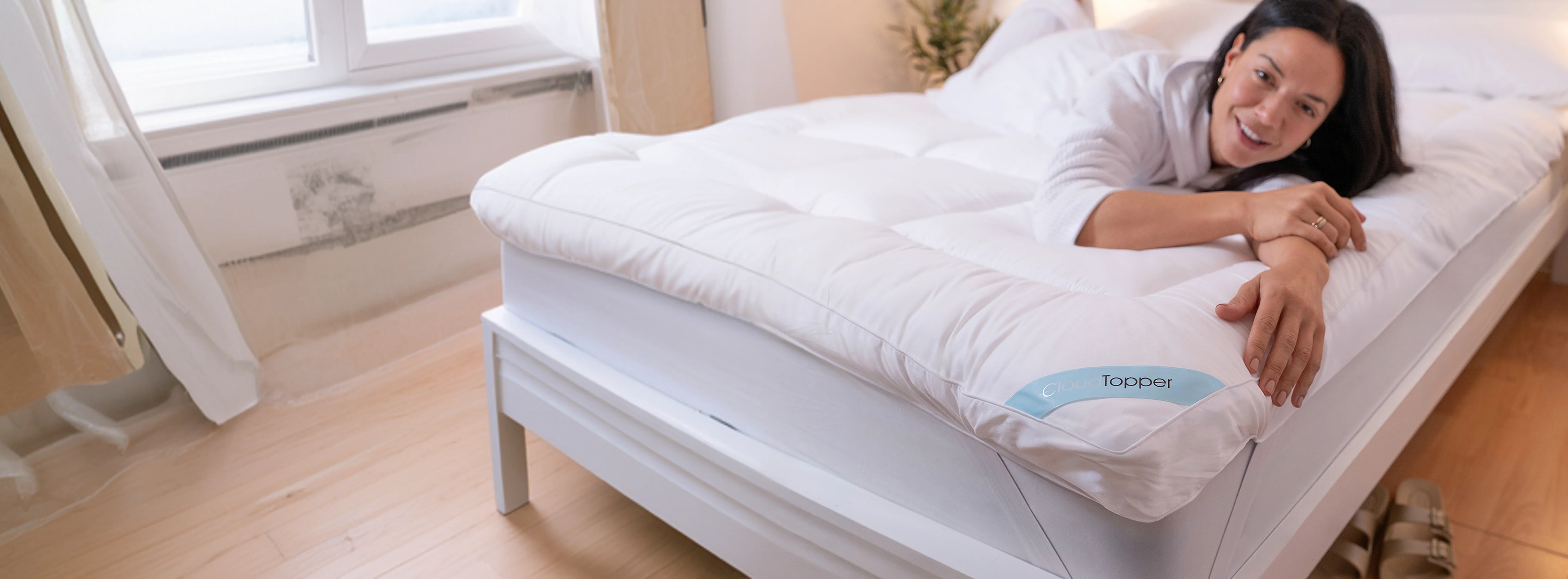 A smiling woman in a white robe lies on her stomach on a bed with a thick mattress topper.