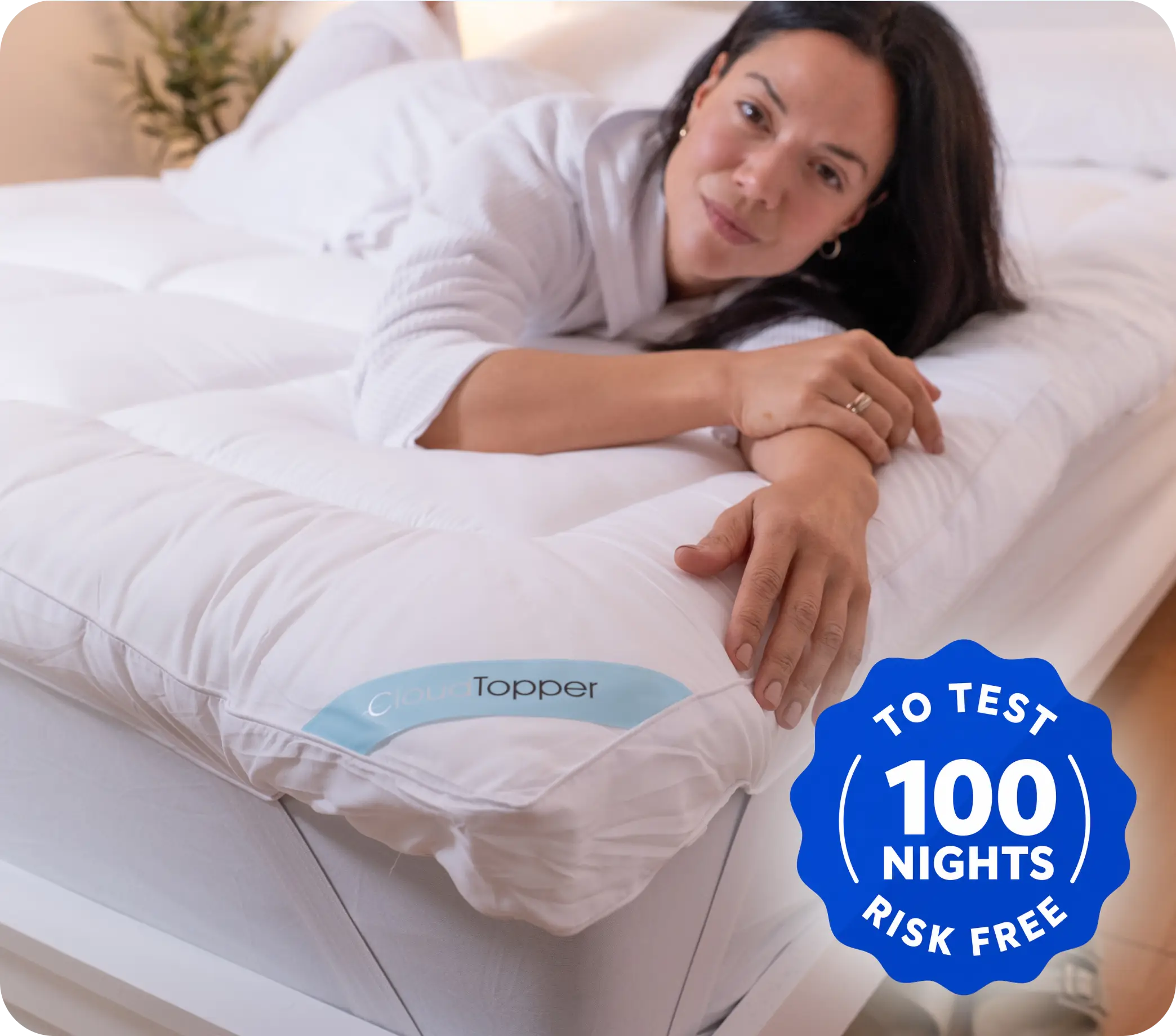 A woman in a white bathrobe lies on her stomach on a bed with a thick mattress topper.