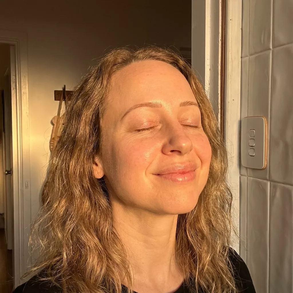 A woman with blonde wavy hair smiles with her eyes closed, her face illuminated by golden sunlight.