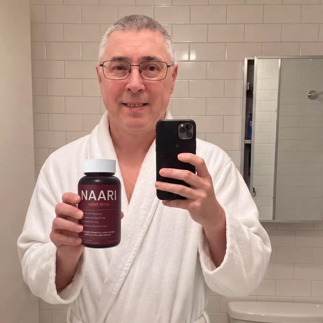 A middle-aged man in a white bathrobe takes a mirror selfie holding a bottle of Naari Heme Iron.
