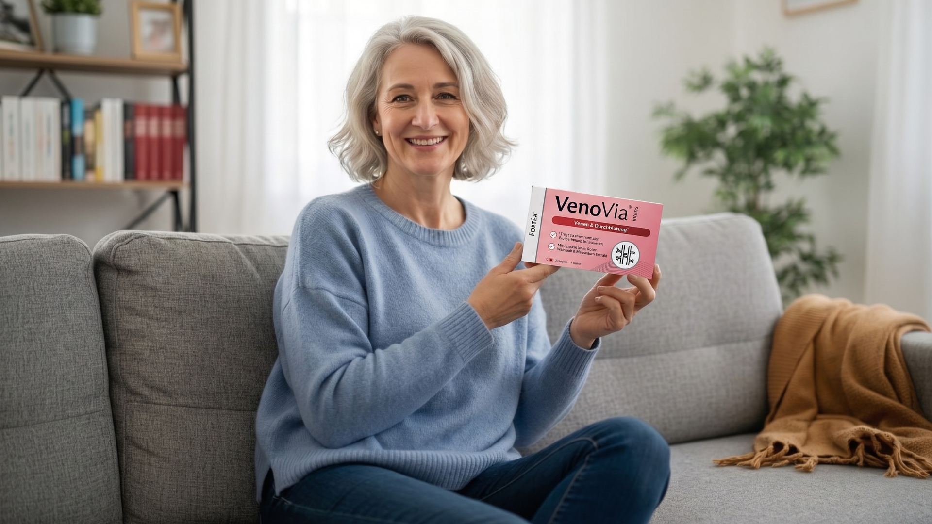 A smiling woman with grey hair sits on a couch, holding a box for an ear product.