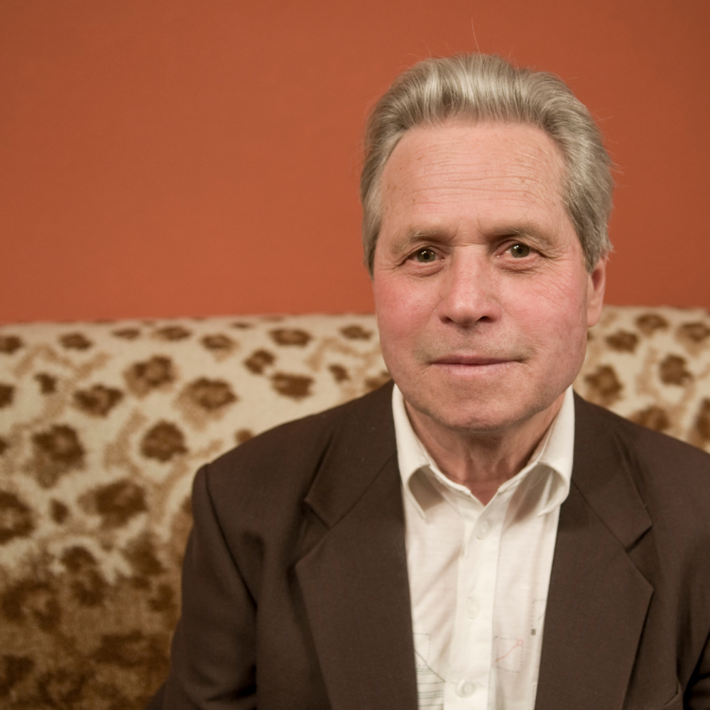 Elderly man in suit jacket sitting on a patterned sofa.