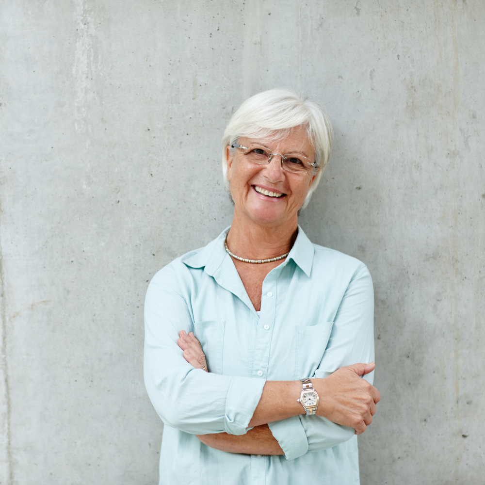 Smiling woman with short white hair, wearing glasses and a light blue shirt, against a concrete wall.