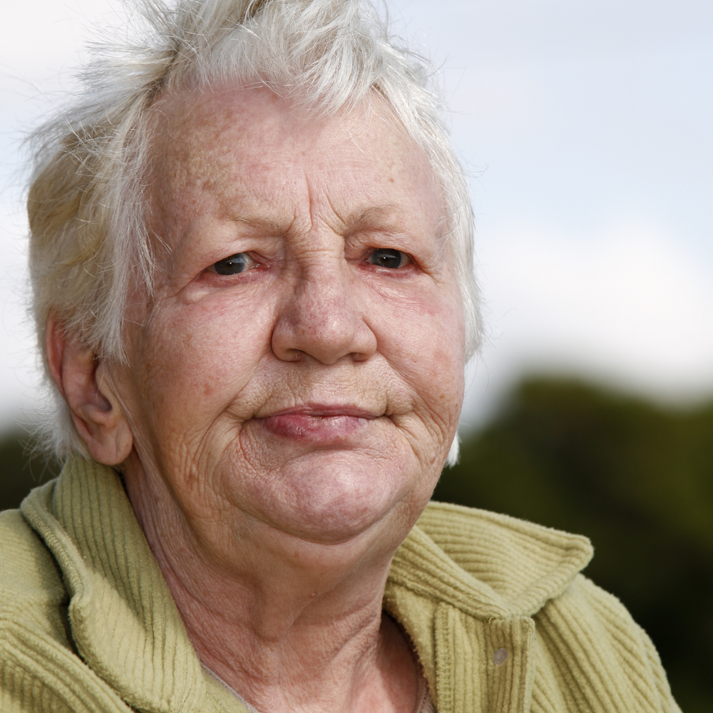 Elderly person with short white hair and a light green jacket outdoors.