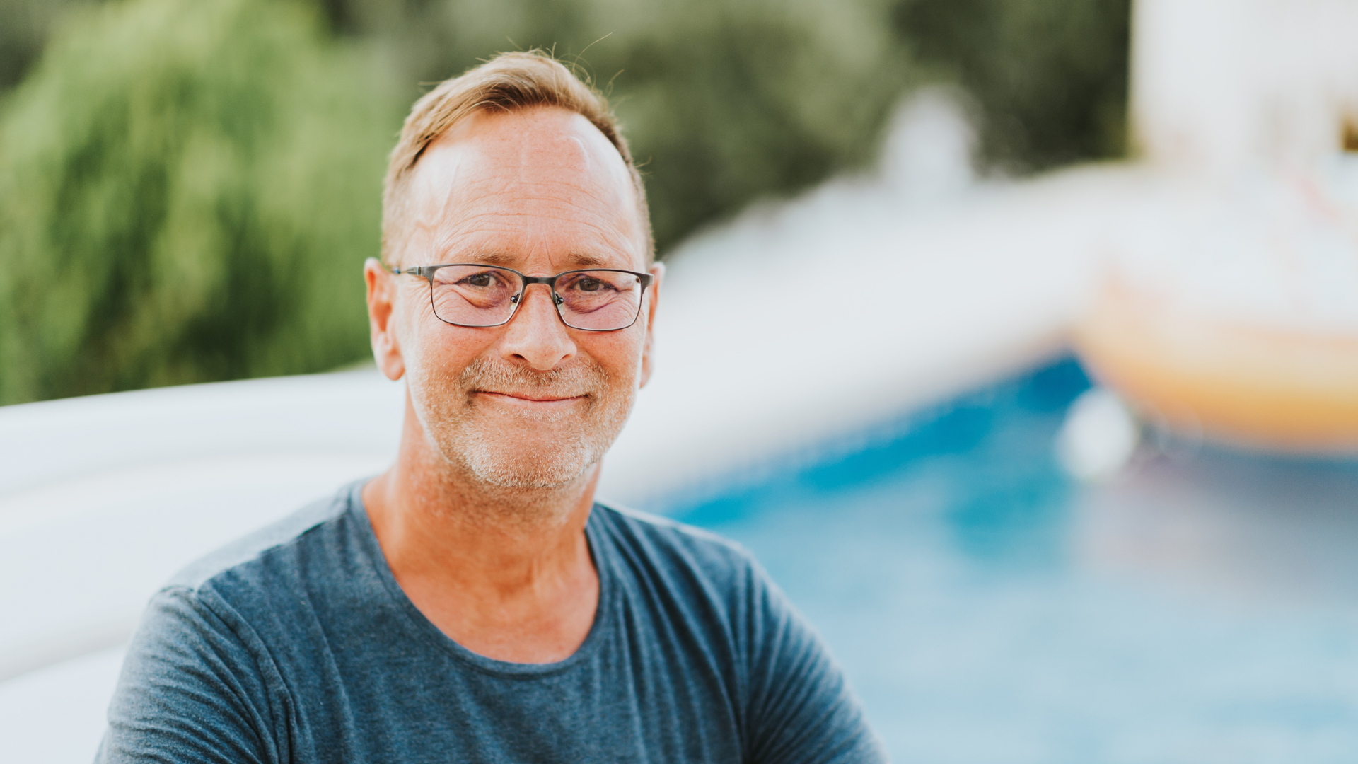 Smiling person with glasses, standing in front of a blurred pool background.