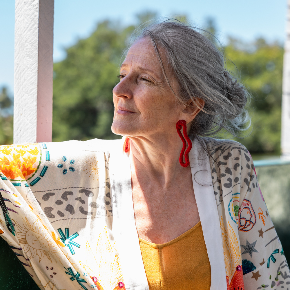 Older woman in colorful outfit standing outside, looking into the distance, with trees in the background.