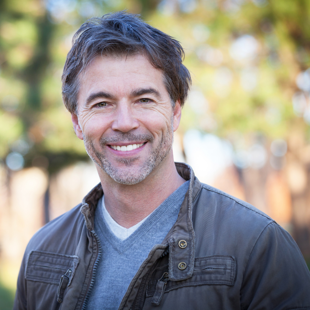 Smiling man with short hair wearing a jacket, standing outdoors with blurred trees in the background.