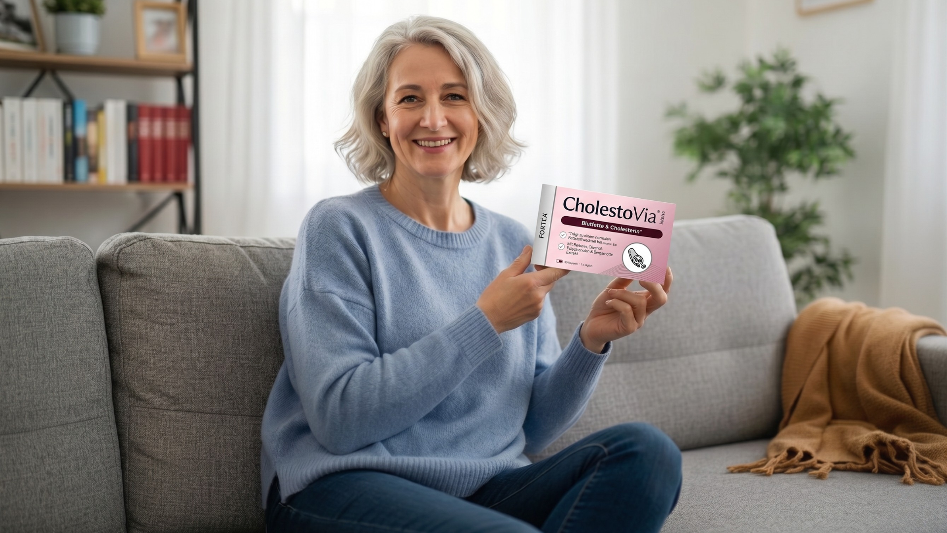 A smiling woman with grey hair sits on a couch, holding a box for an ear product.