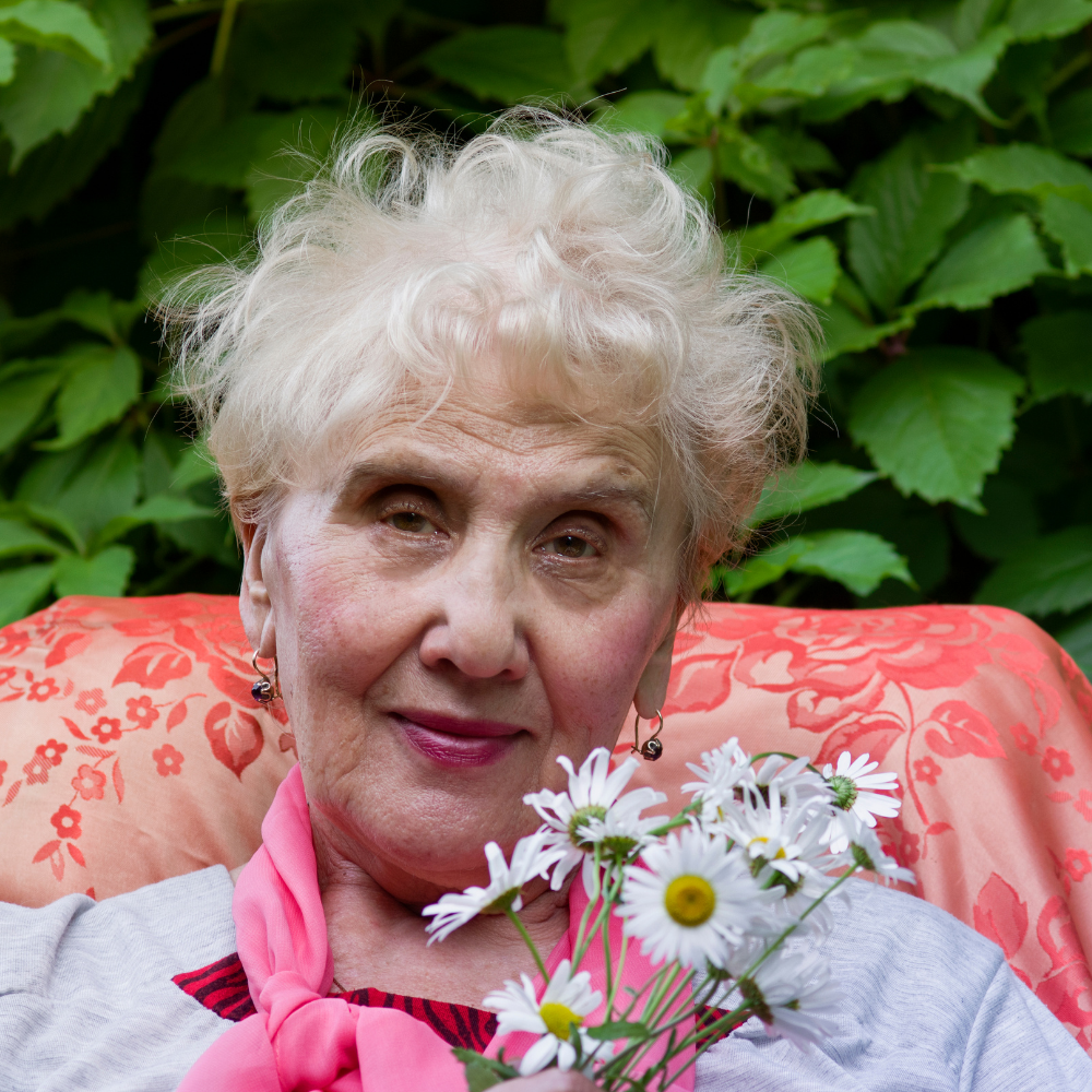 Woman with white hair holding daisies, seated outdoors with green plants in the background.