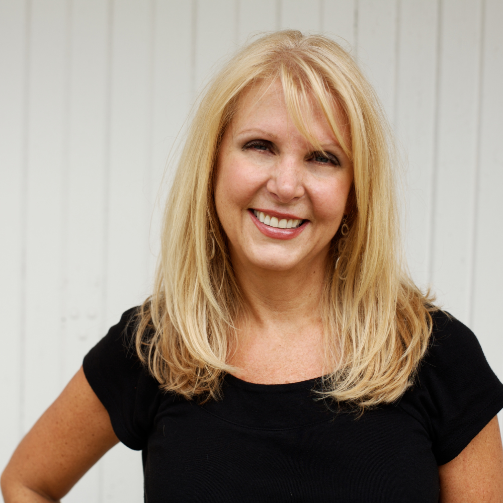 Smiling person with long blonde hair in front of a white wooden background.