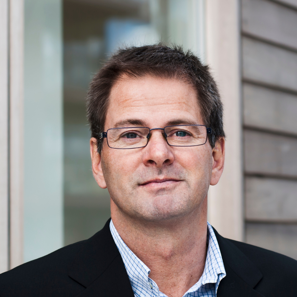 Man in glasses wearing a suit, standing in front of a blurred background.