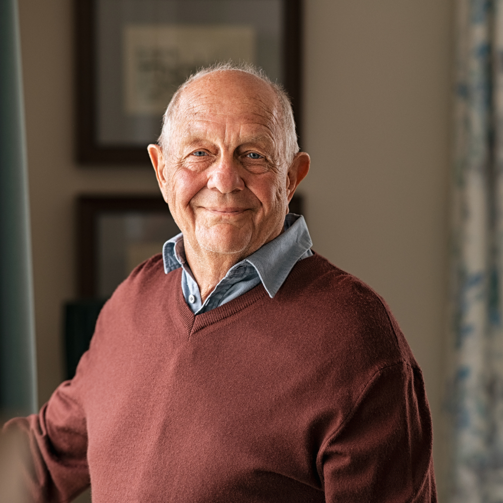 Smiling elderly man in a maroon sweater stands in a room with framed pictures.