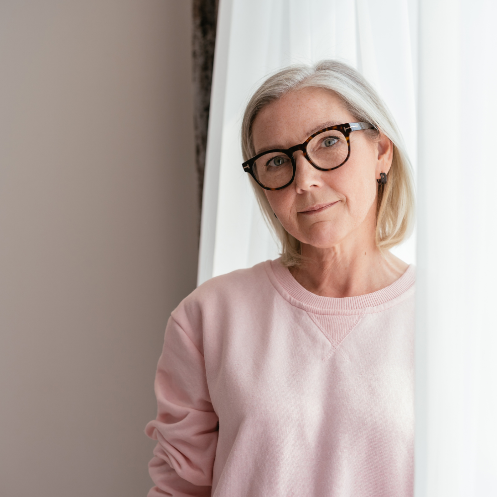 Person with glasses and white hair wearing a pink sweater, standing by a curtain.