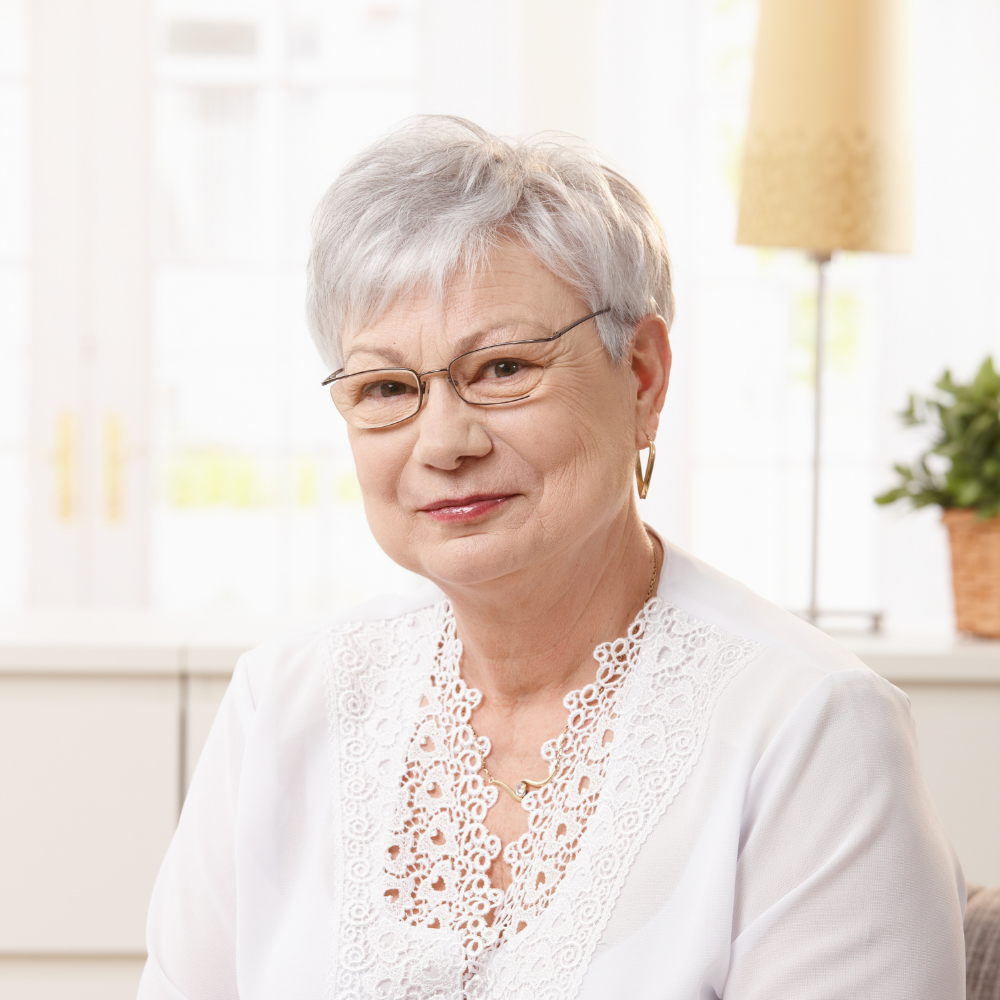 Elderly woman with short silver hair and glasses, wearing a white blouse, smiling indoors.