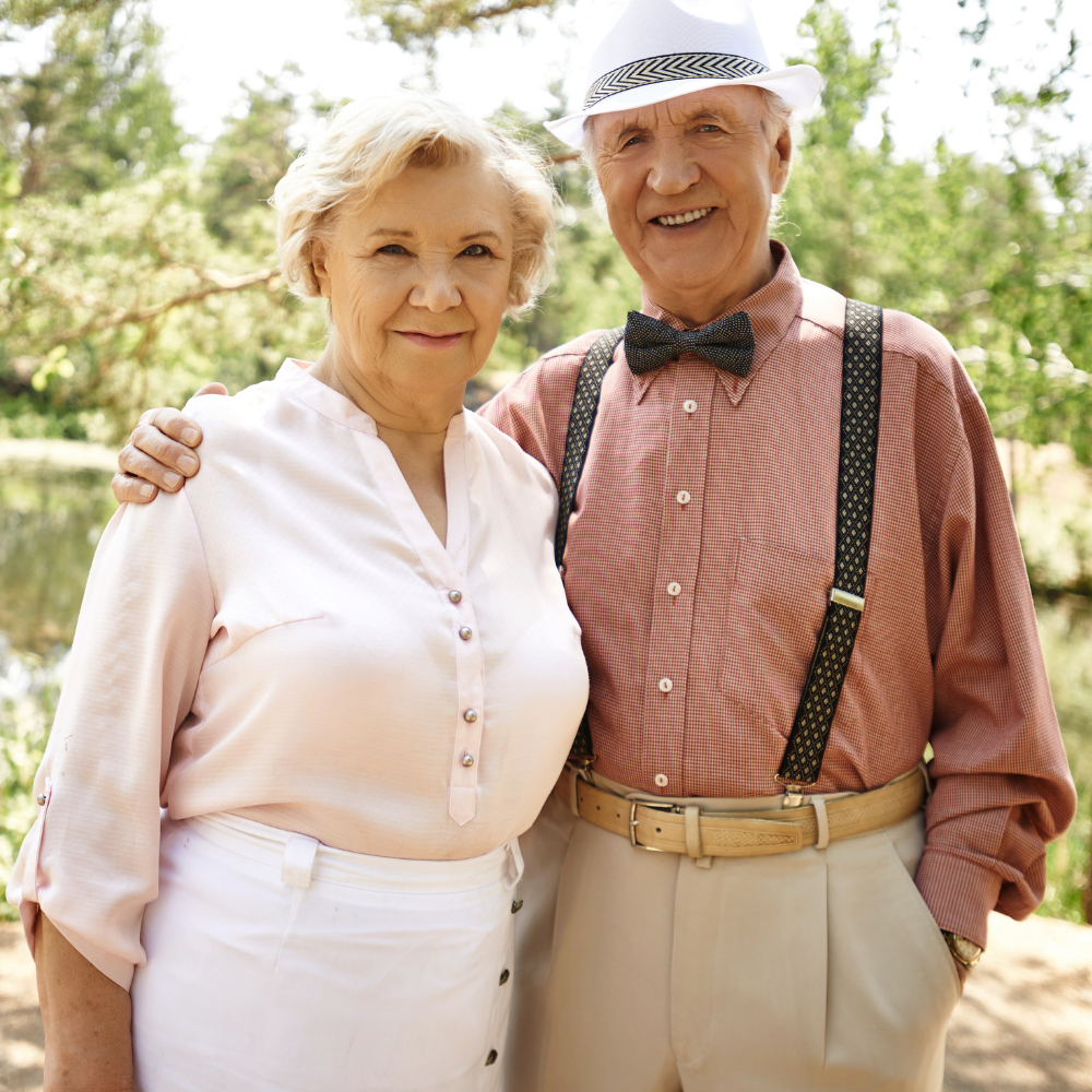 Elderly couple smiling outdoors, man in hat and bow tie, woman in light blouse.