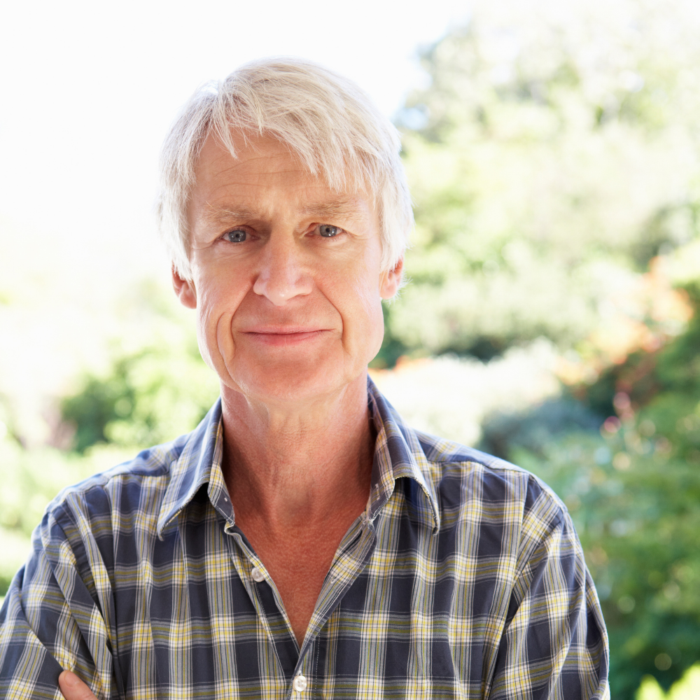 Older man in a plaid shirt standing outdoors with greenery in the background.