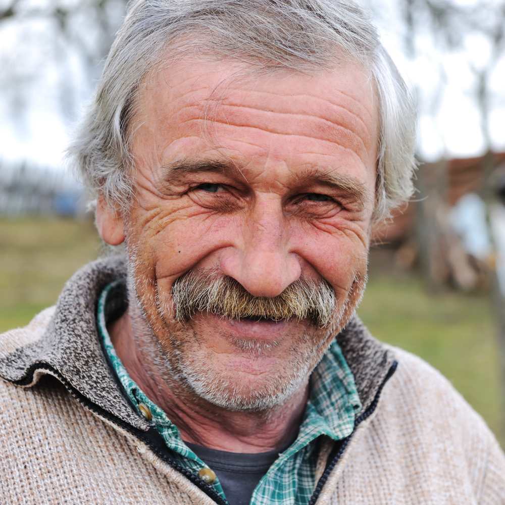 Smiling older man with gray hair and a mustache, wearing a plaid shirt and sweater.