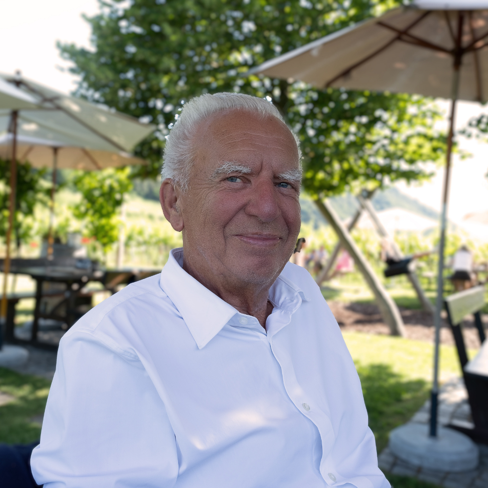 Older man smiling outdoors, sitting under an umbrella in a garden setting.