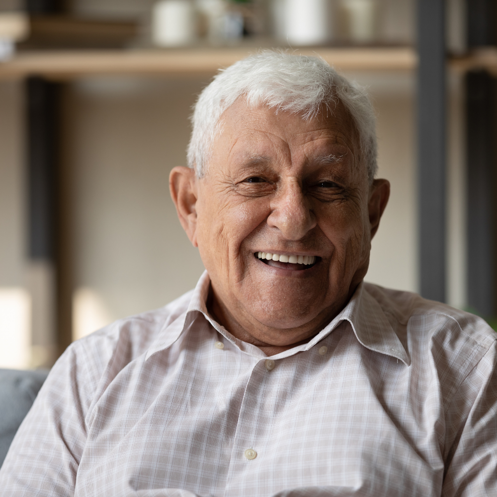 Elderly man smiling indoors, wearing a light-colored shirt.