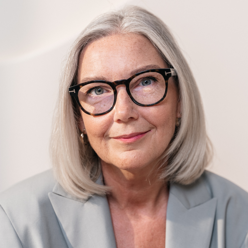 Woman with gray hair and glasses wearing a light gray blazer.
