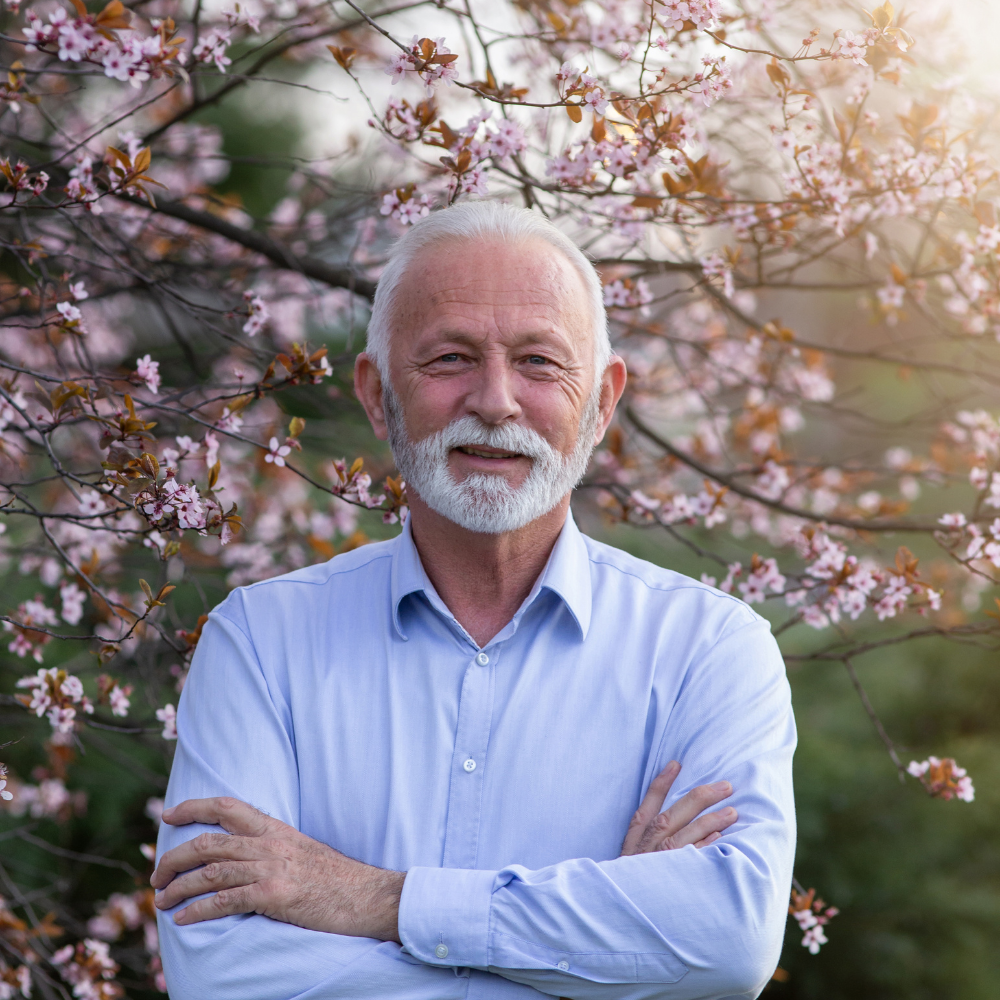 Older man with a beard, standing among blooming cherry blossoms.