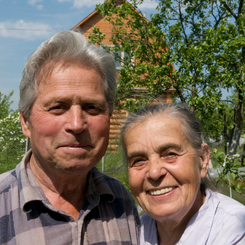Smiling elderly couple standing outside near trees and a wooden house.
