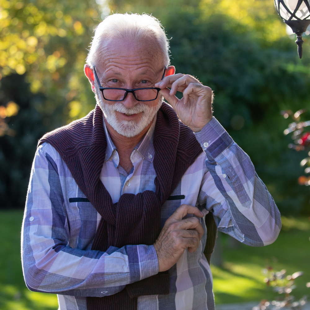 Older man adjusting glasses outdoors with trees in the background.