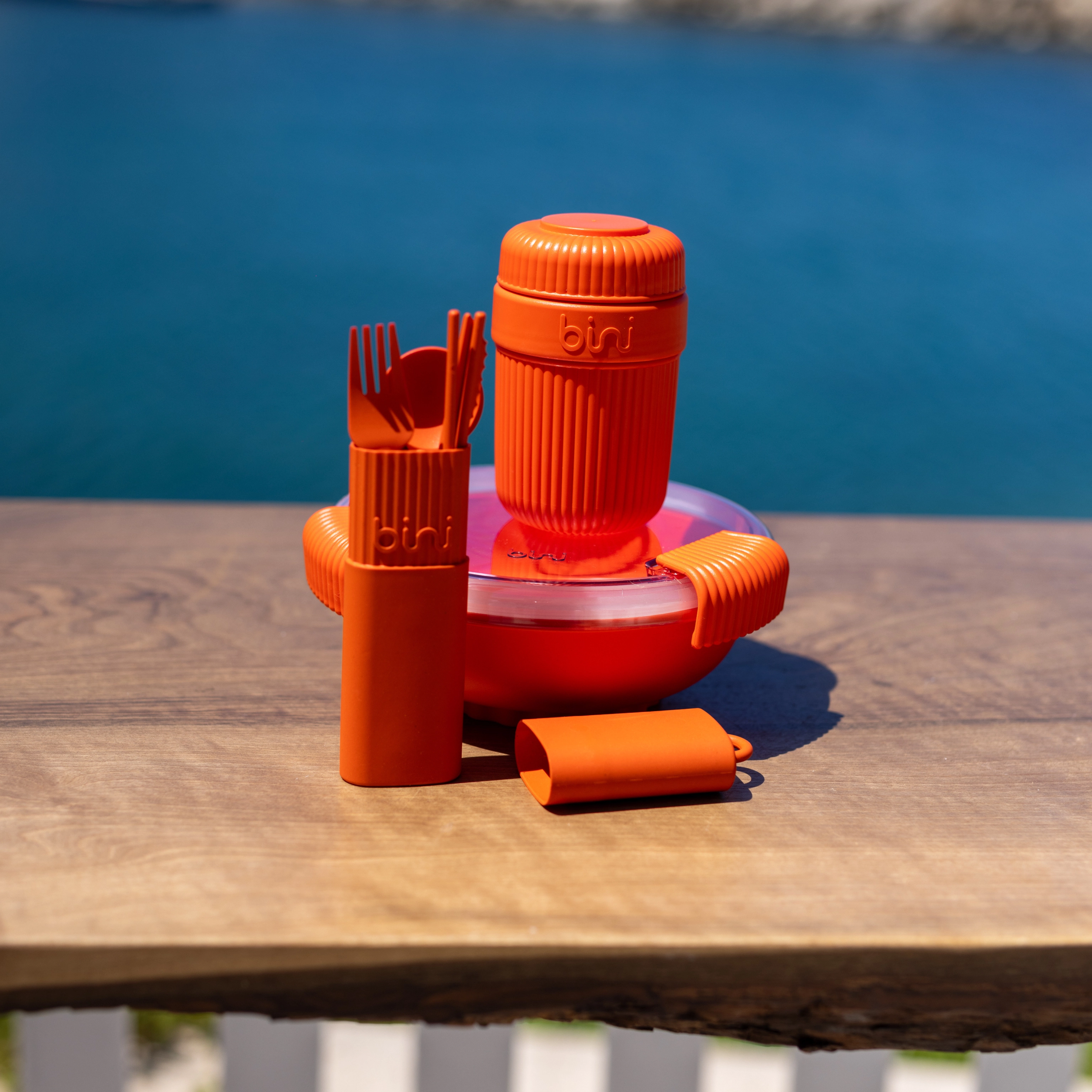 An orange Bini reusable lunch set, including a bowl and cup, on a wooden surface outdoors.
