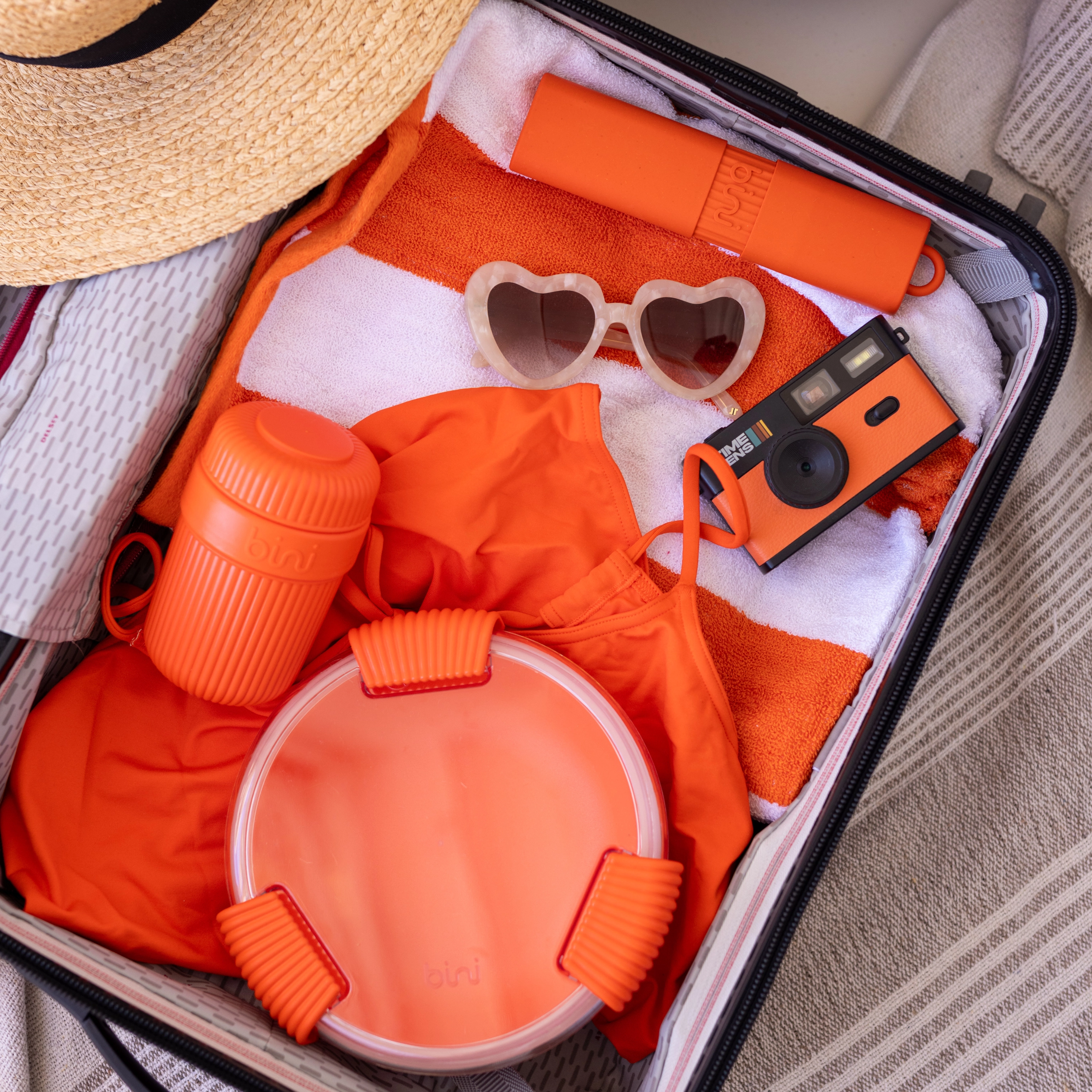 An open suitcase packed with orange beach items including a swimsuit, towel, camera, and heart-shaped sunglasses.