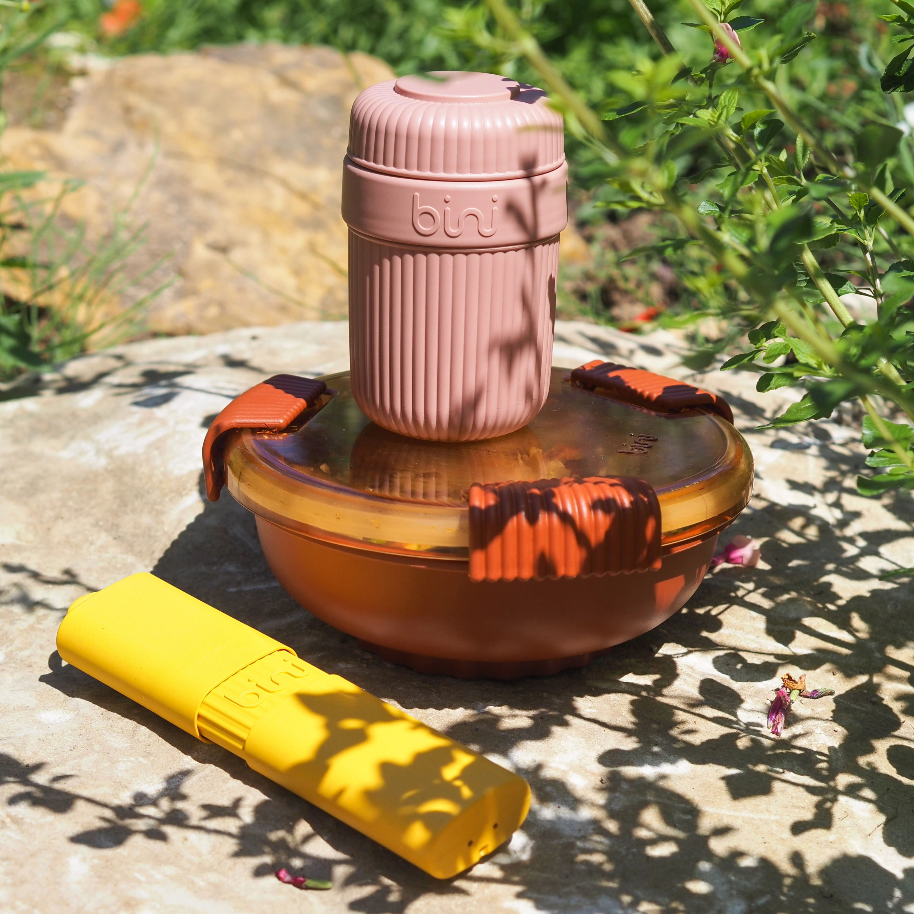 A pink travel cup, brown food container, and yellow case sit on a rock in the sun.