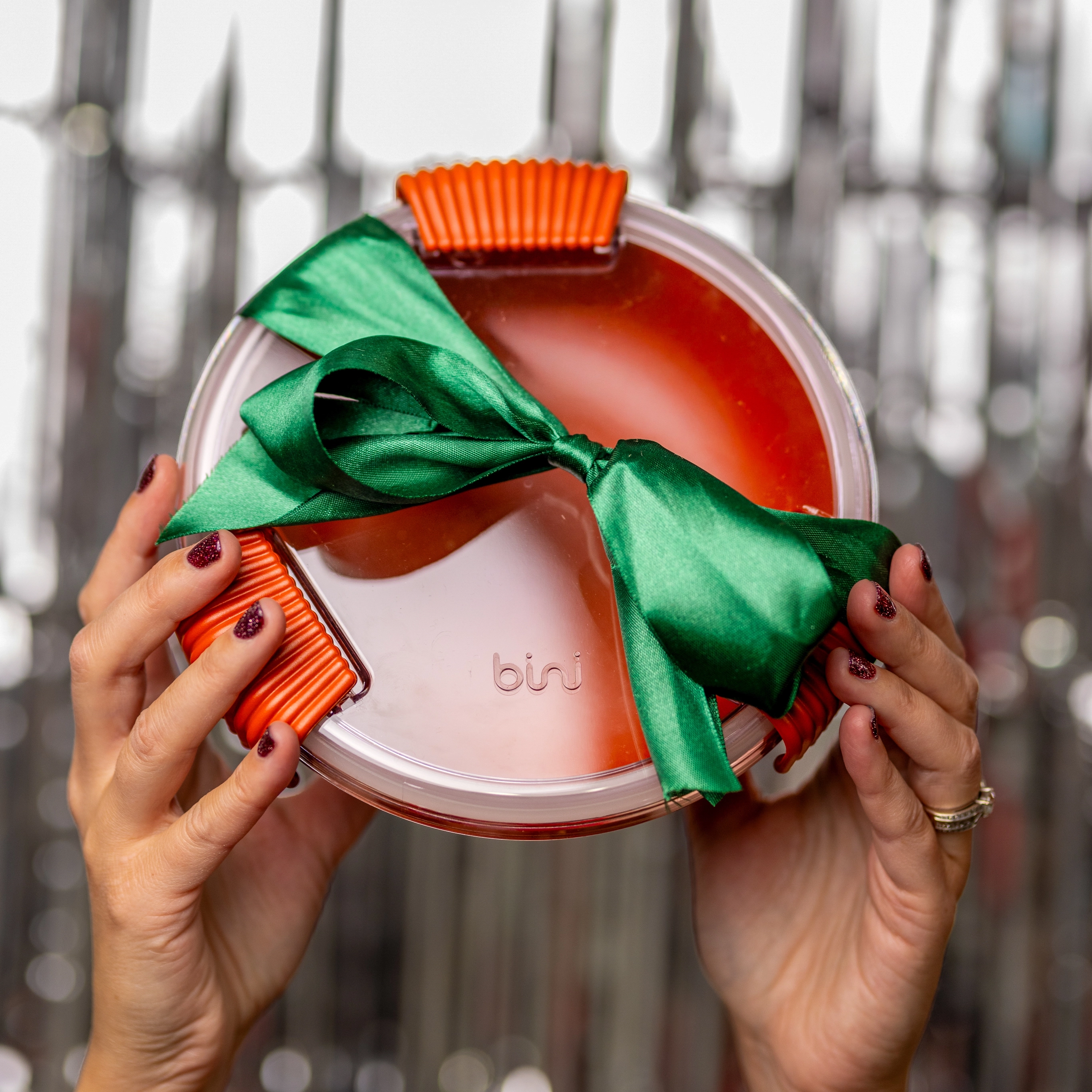 Hands holding a round container with orange accents and a green ribbon.