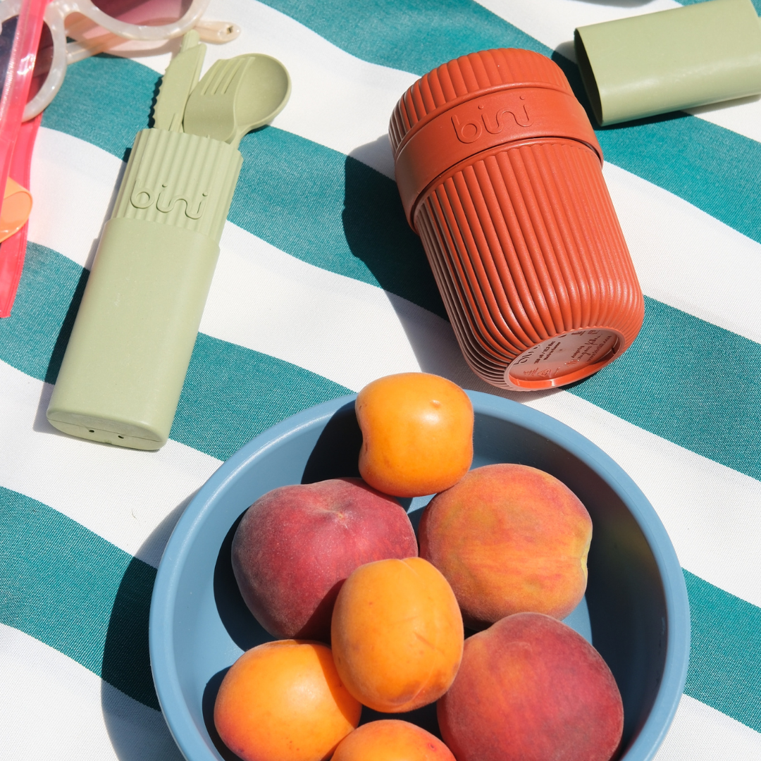 Peaches and apricots in a bowl with reusable utensils and cup on striped fabric.