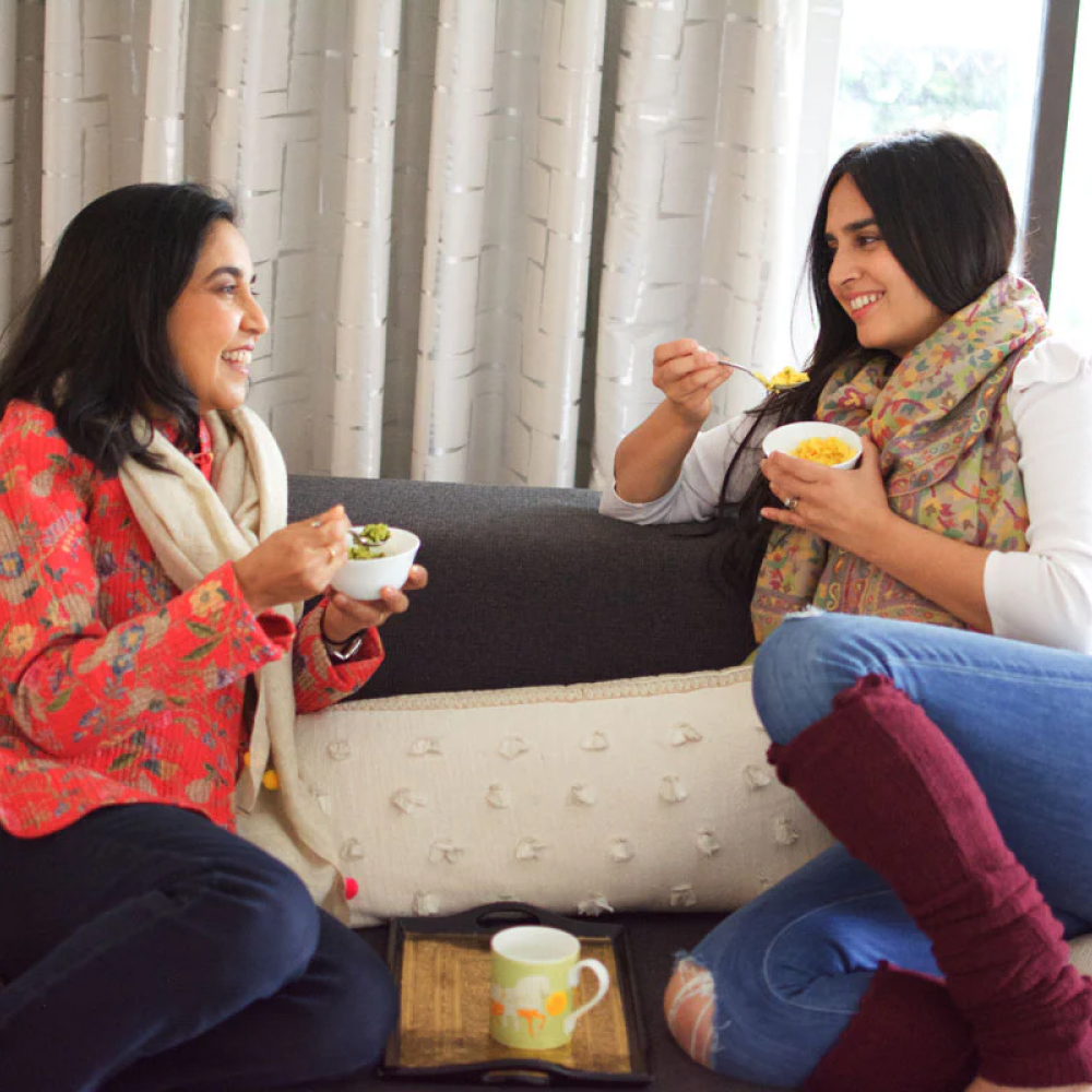 Two women sitting on a couch, eating from bowls and smiling at each other.