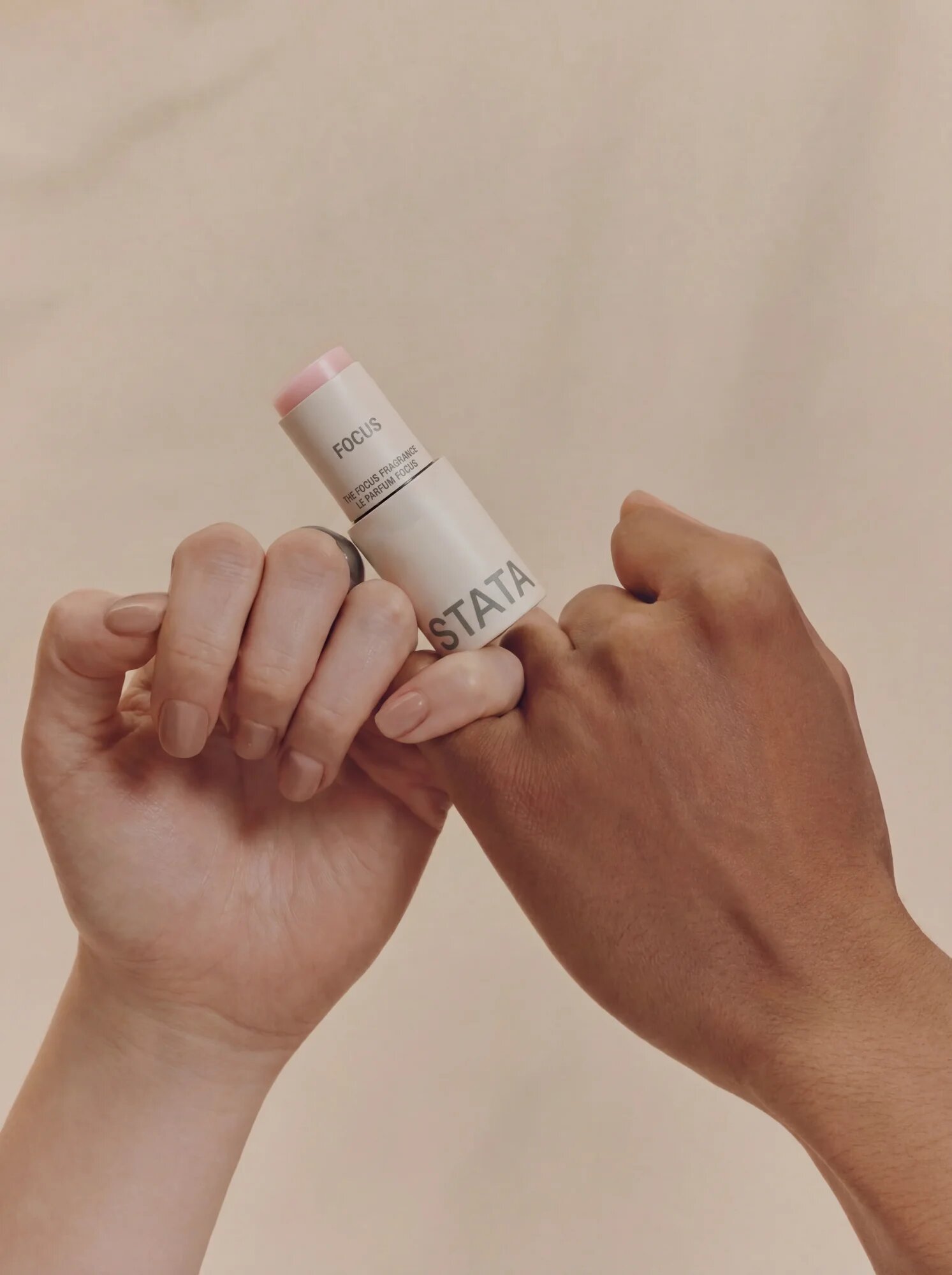 Two interlocked hands of different skin tones hold a white cosmetic stick against a plain background.