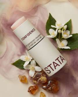 A white tube of solid perfume labeled 'Confidence' and 'Stata,' surrounded by jasmine flowers and amber resin.